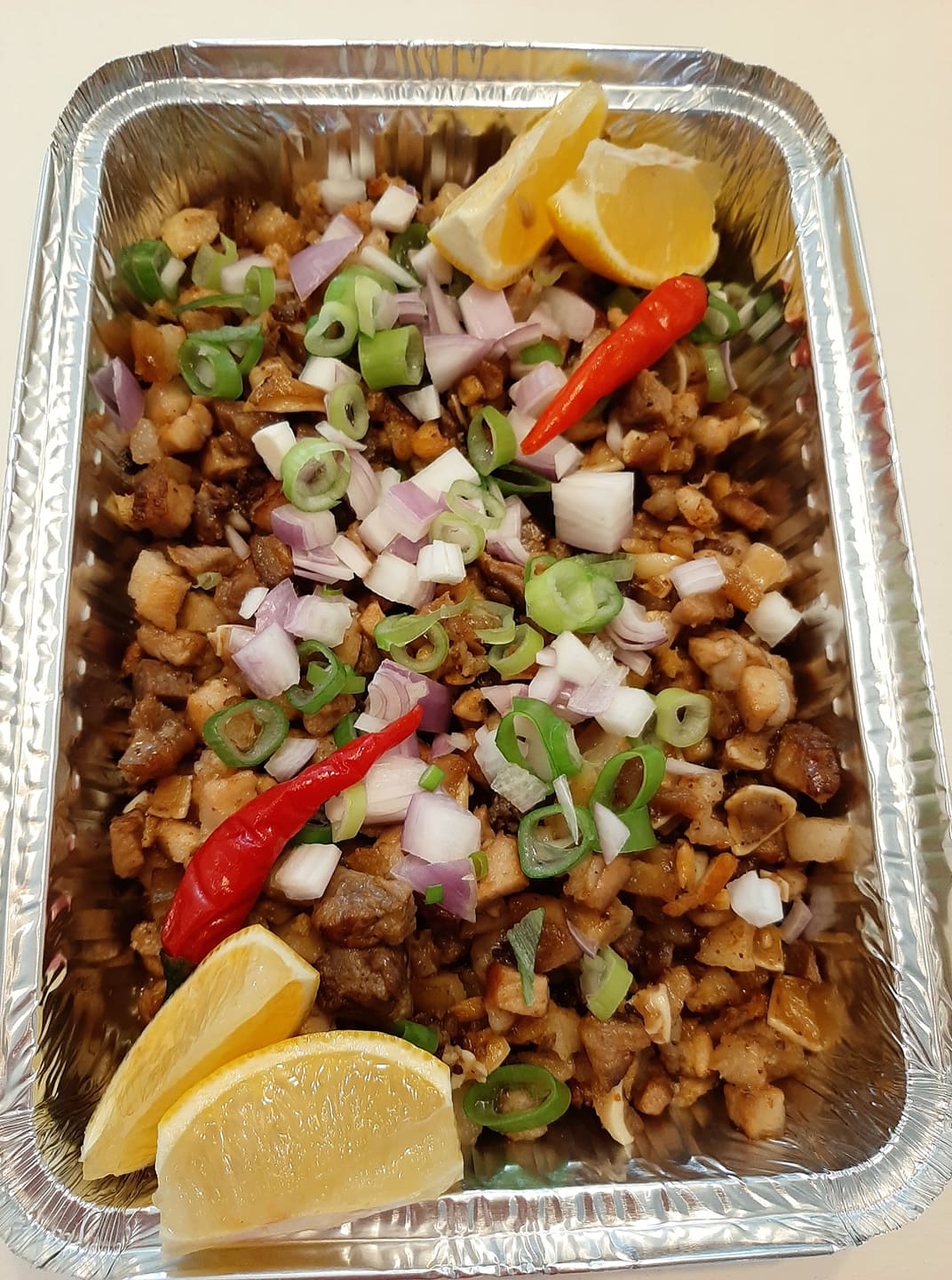 Pork sisig in a foil tray garnished with chillies, spring onions and lemon