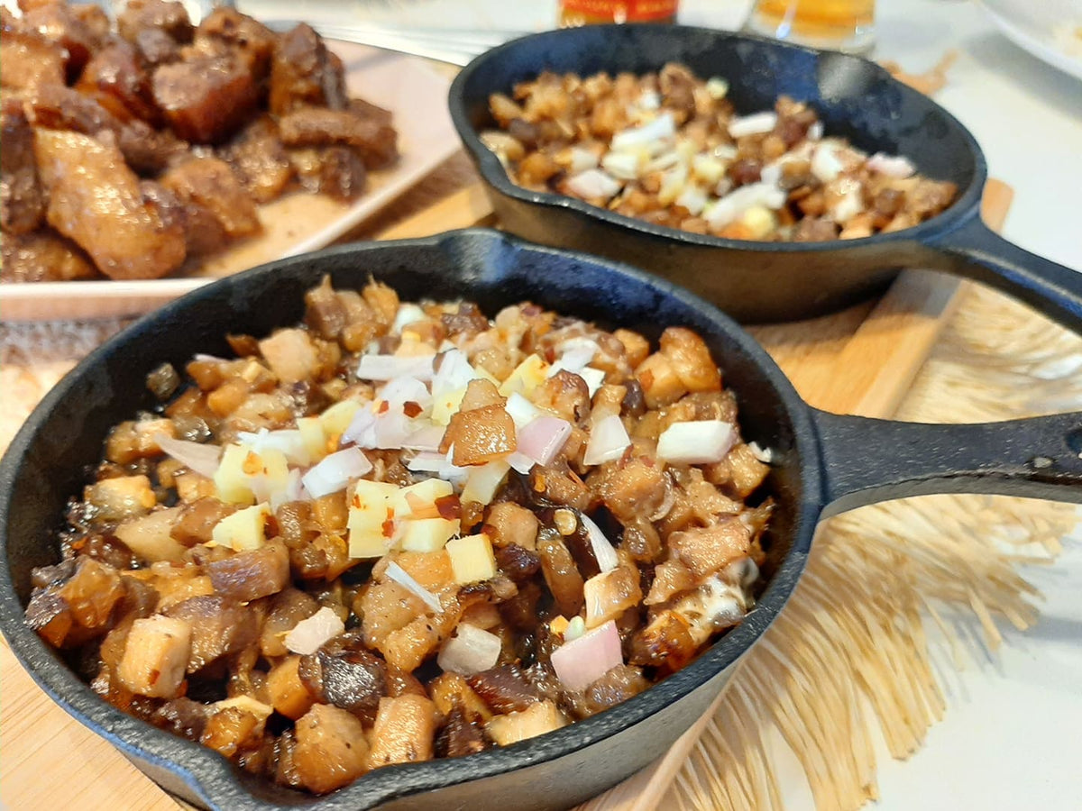 Two sizzling skillets of pork sisig topped with chopped onions